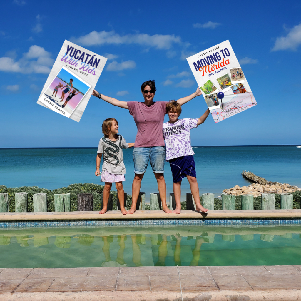 Cassie holding copies of giant books in each hand. Kid on either side. Books are: Yucatan with Kids and Moving to Merida. Standing in front of a pool, blue ocean and sky behind