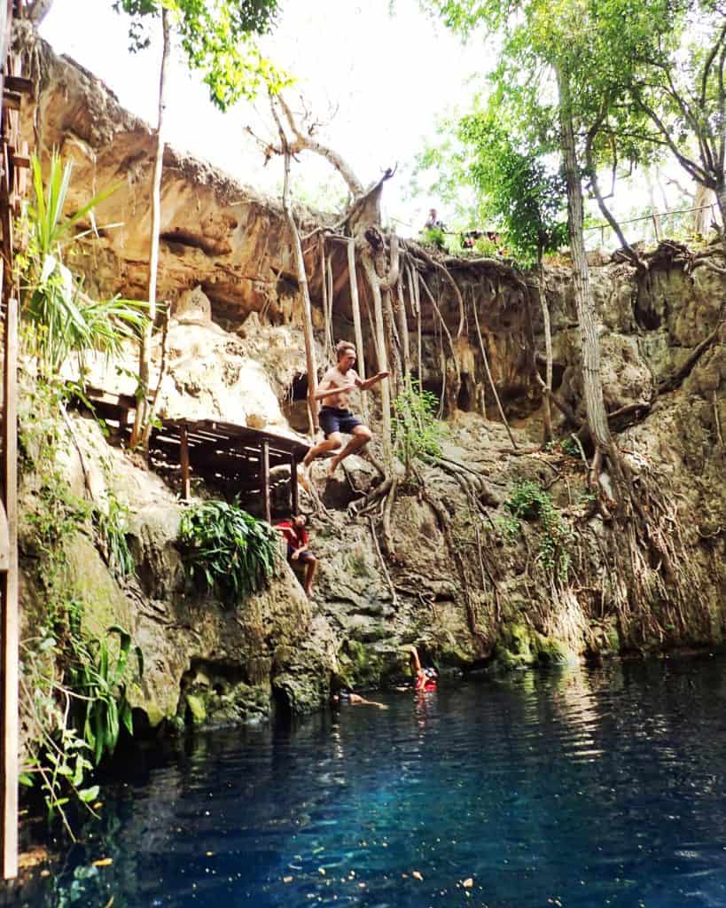 man jumping off a wooden platform into a cenote. blue water, trees all around