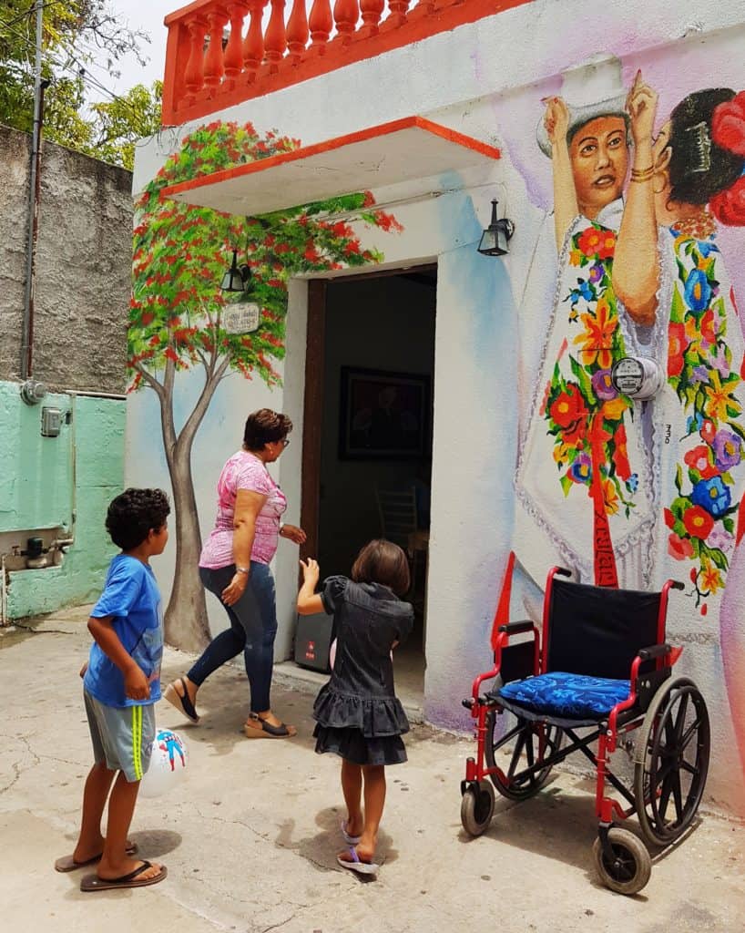 wall with two people painted on, both in traditional Maya dress. Kids in front playing and a wheelchair in the road
