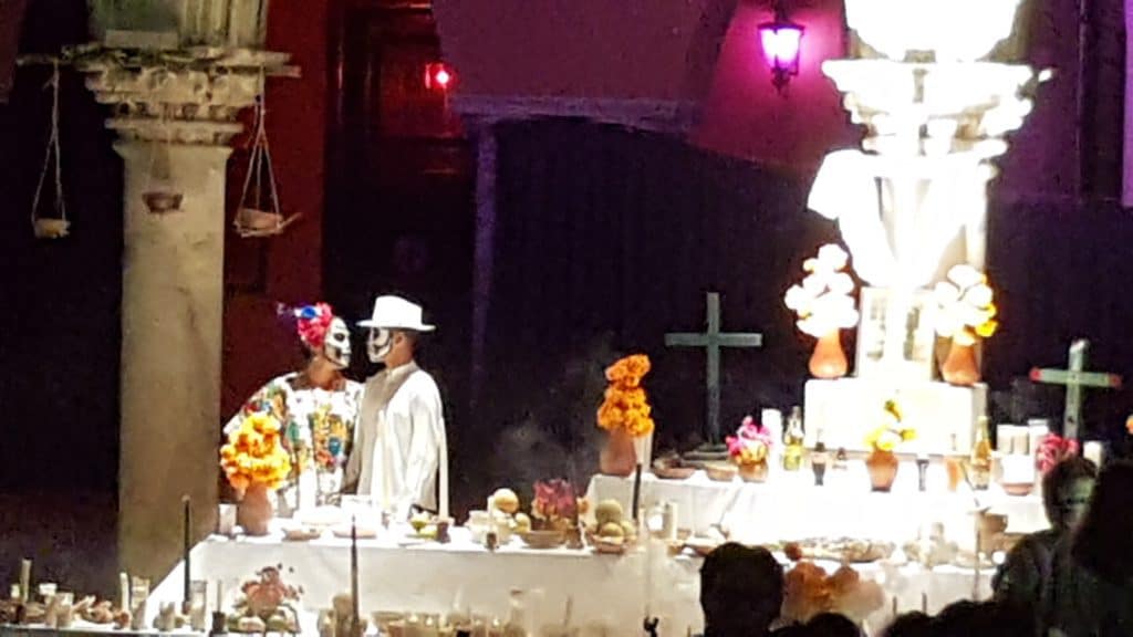 day of the dead dance on plaza in merida - man and woman in traditional white clothes with faces painted stand behind a table altar