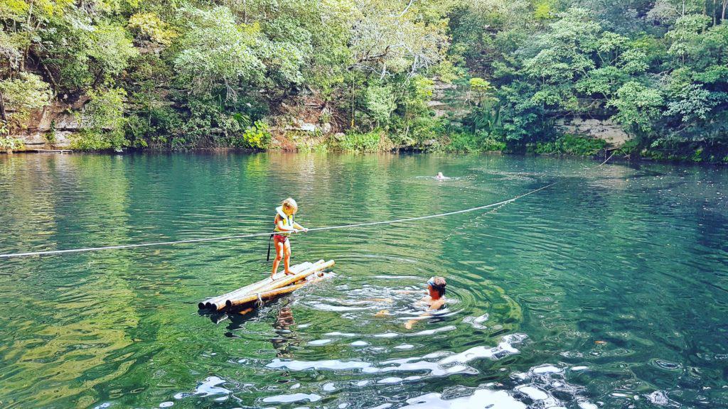little kid standing on bamboo raft in middle of large cenote. Green water, trees all around