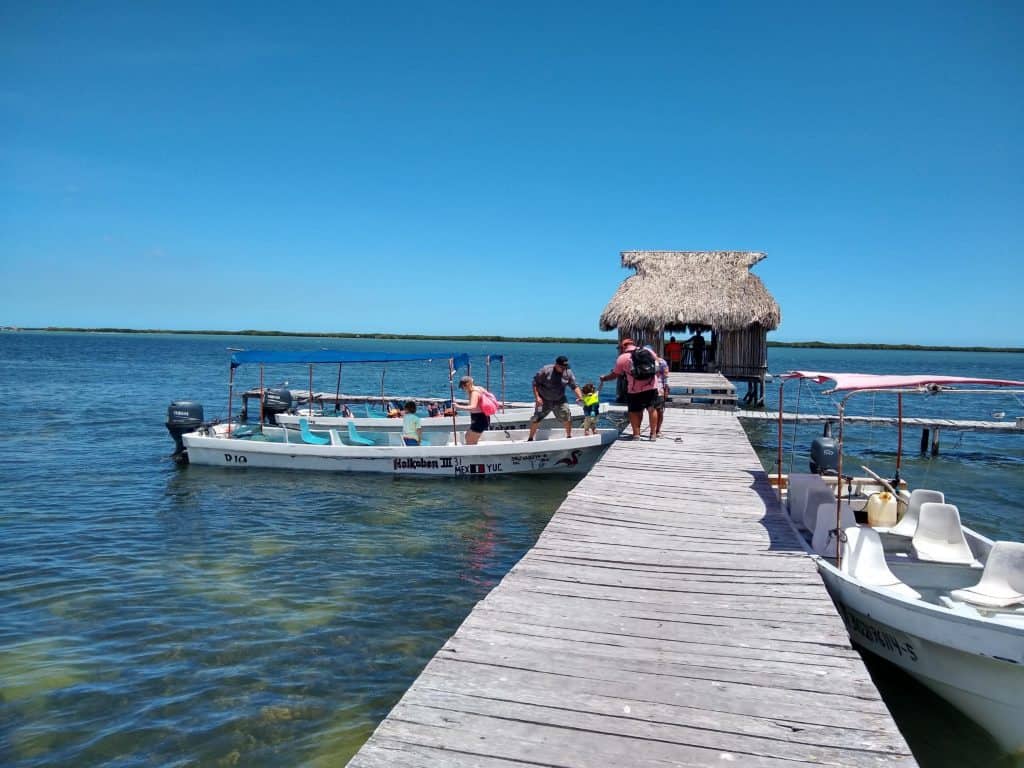 Small wooden jetty on blue sea. Thatch at end, blue sky. People getting on a small boat