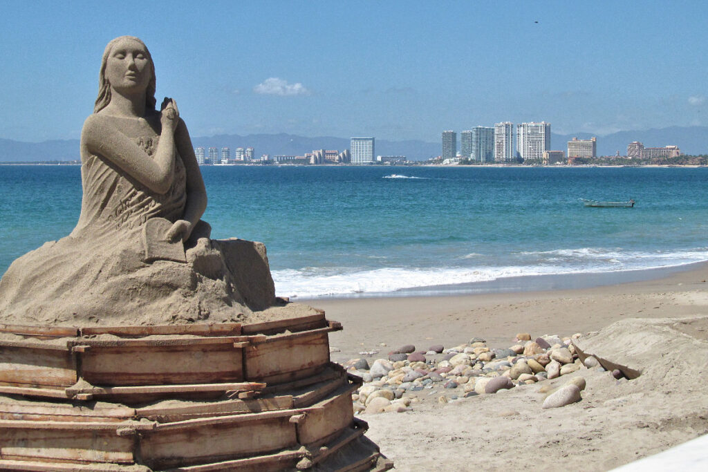 view of a sand statue (of a woman) on Puerto Vallarta city beach. Highrise buildings on other siide of bay