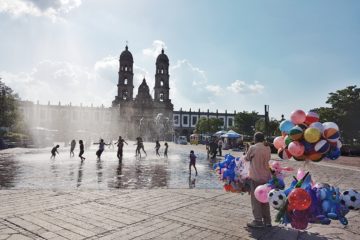 convent in Zapopan, Jalisco behind water fountain and kids playing. Man with kids toys for sale in right corner of picture