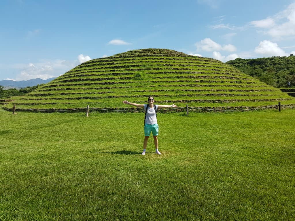 round, stepped pyramid covered in grass. Woman in green shorts, arms outstretched in front of it