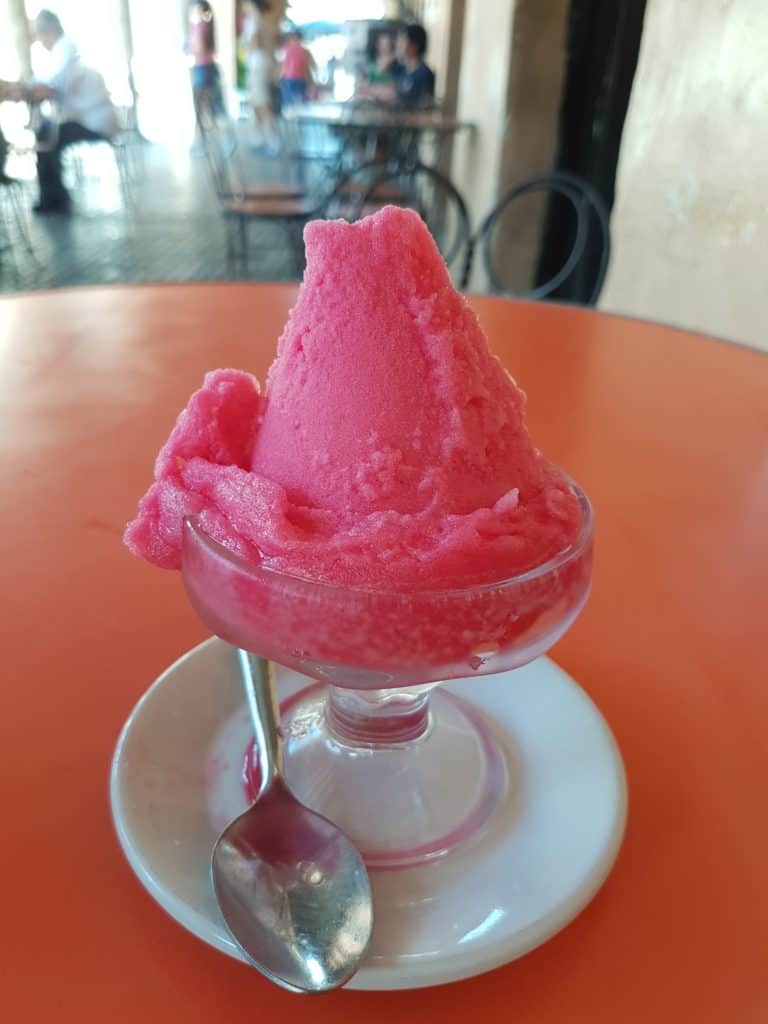 watermelon ice cream in a glass bowl n a white saucer with spoon. Red table.