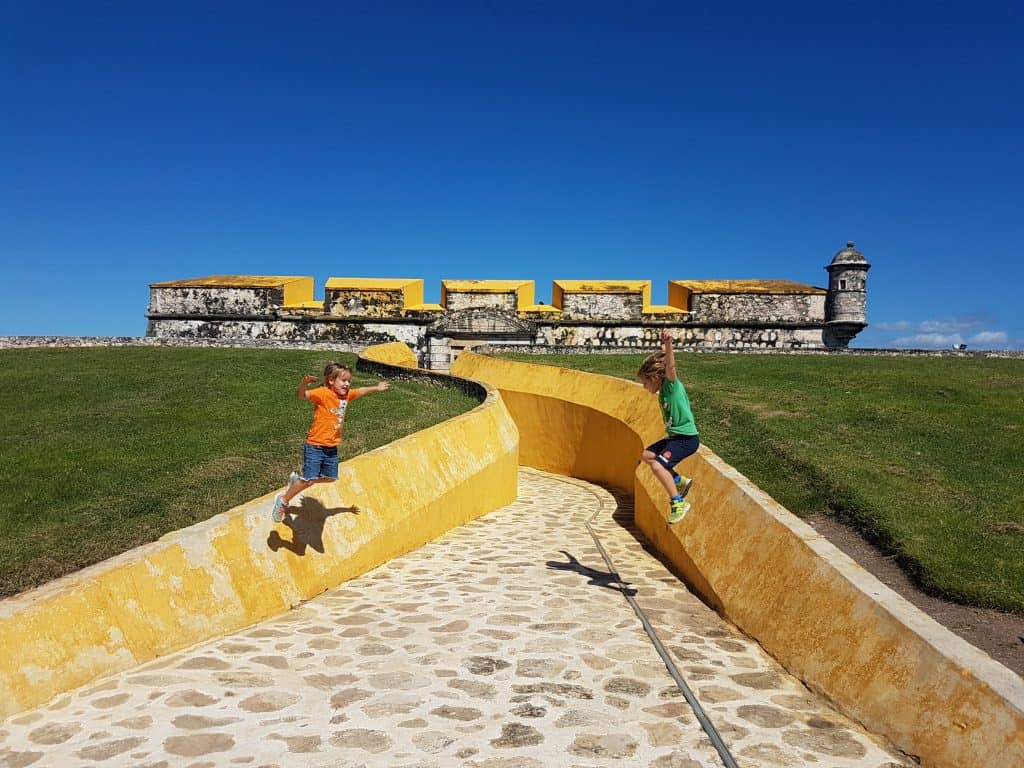 children jumping off yellow walls. one in orange, one in green. fort top in background. grass and blue sky