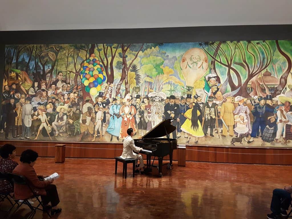 Man playing grand piano infront of Diego Rivera mural