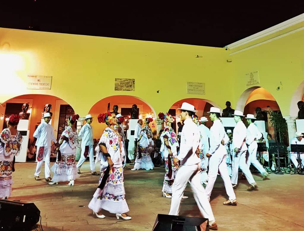men and women in traditional yucatecan dress dancing on stage, yellow walls behind