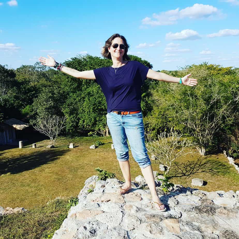 Cassie standing on top of a ruin, arms out, wearing short jeans and a blue shirt