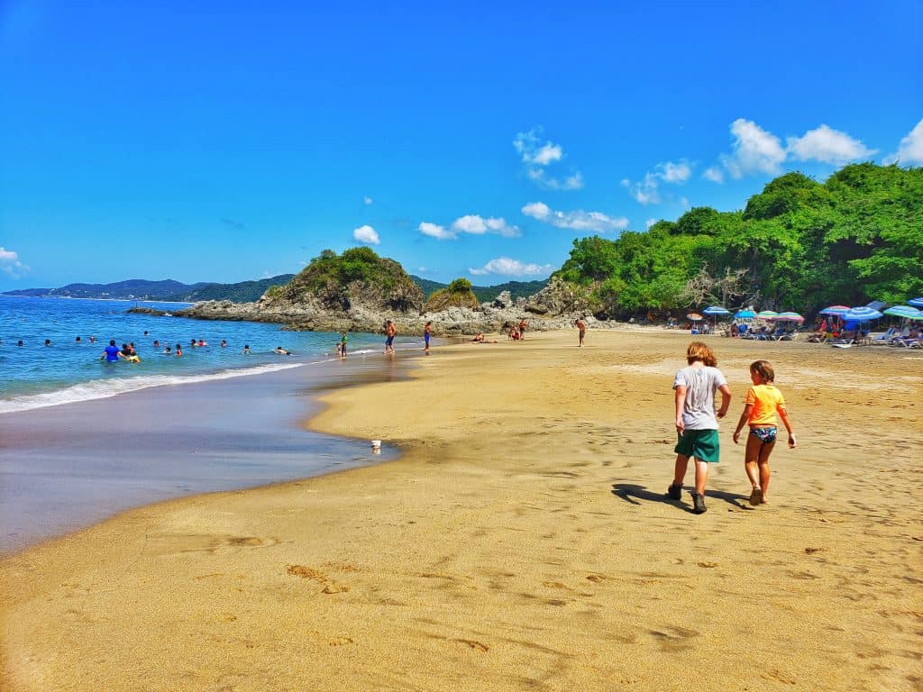 two small children walk along a beautiful sandy beach. Blue sea, trees and rocks in distance, blue sky