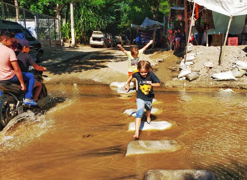 two small children running over sandbag stepping stones in a shallow brown watered river
