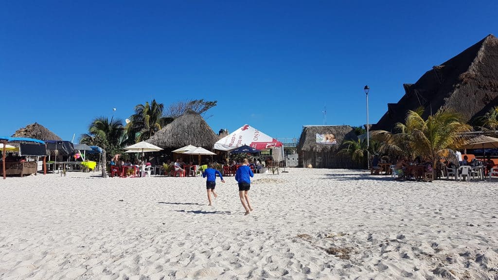 two kids in blue shirts and black shorts running towards tables with umbrellas on the beach. blue sky