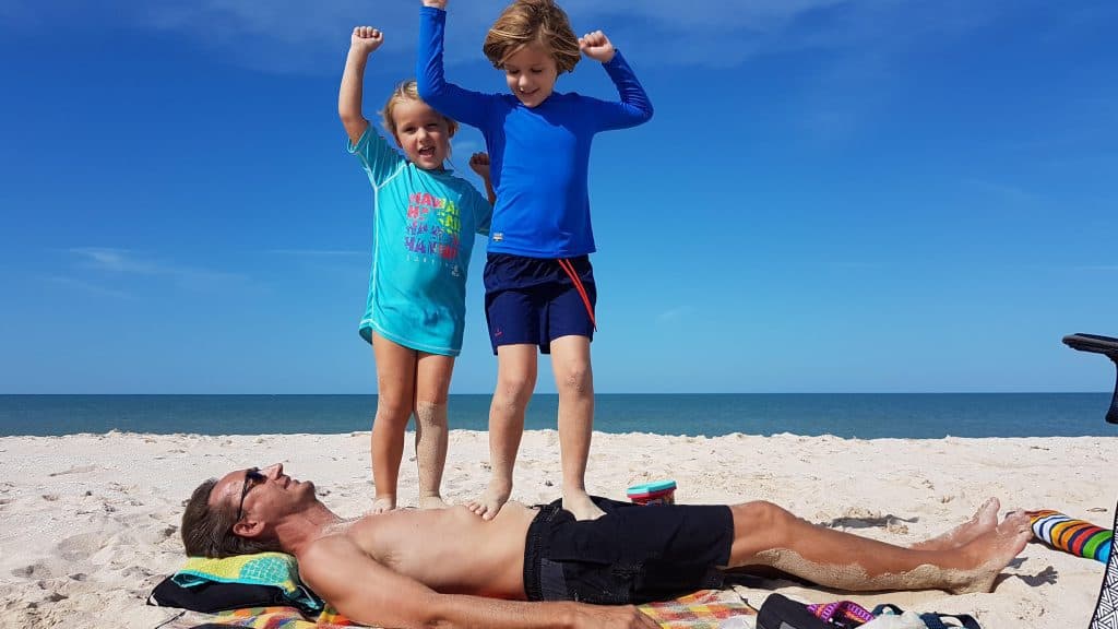 two small kids standing on their dad while he lies on the beach