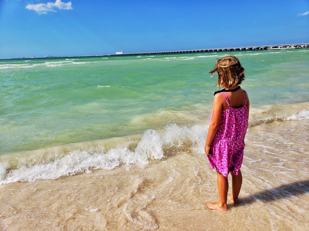 little girl in pink standing in front of clear green sea, blue sky
