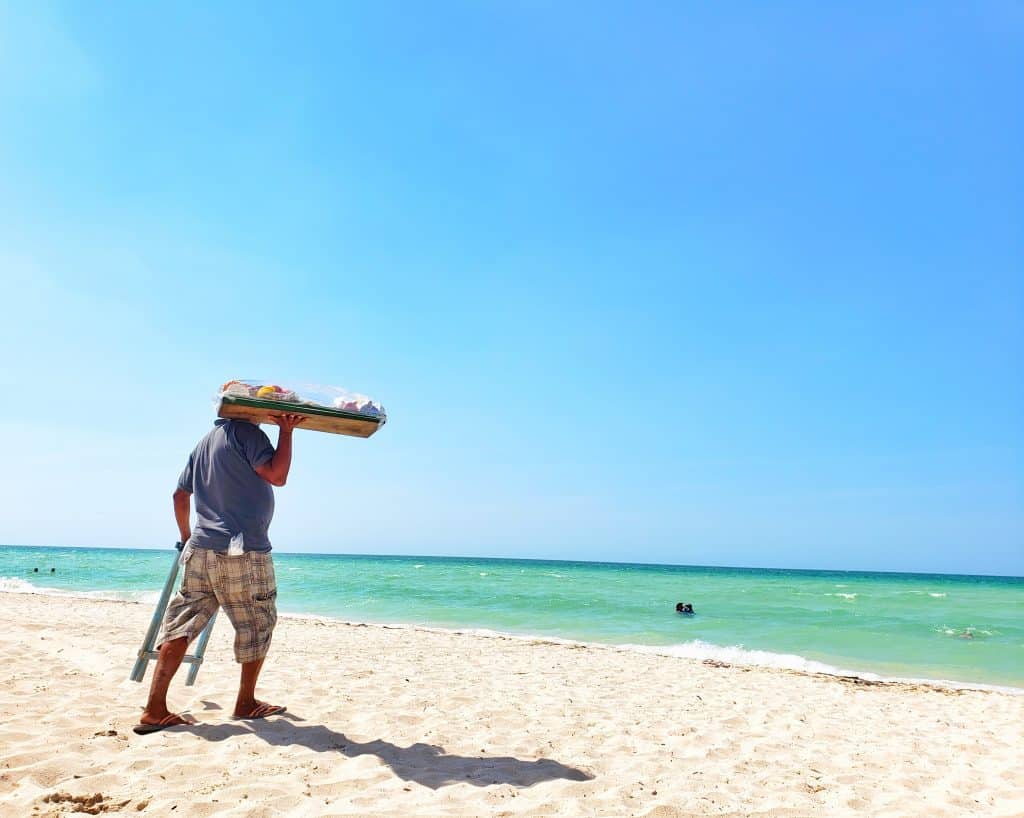 snack seller walking on sand, green sea, blue sky. he holds a wooden tray of candies