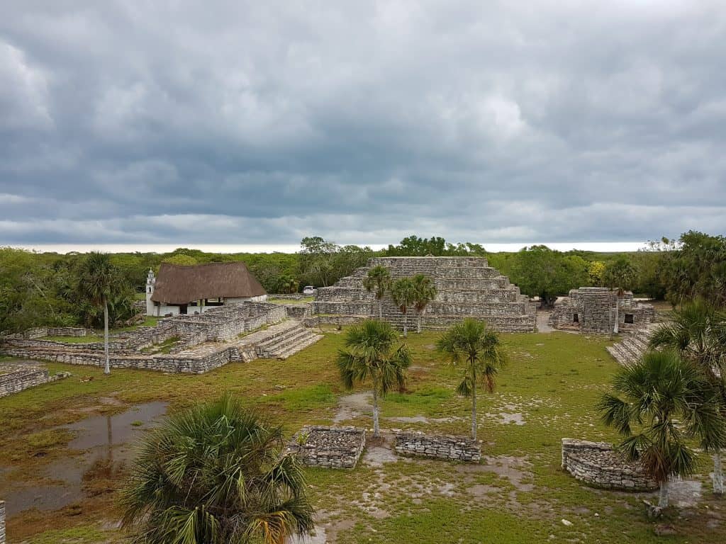 view of x'cambo from top  of the pyramid. thatched chapel on left, pyramid in front, low  ruins on ground. grey sky