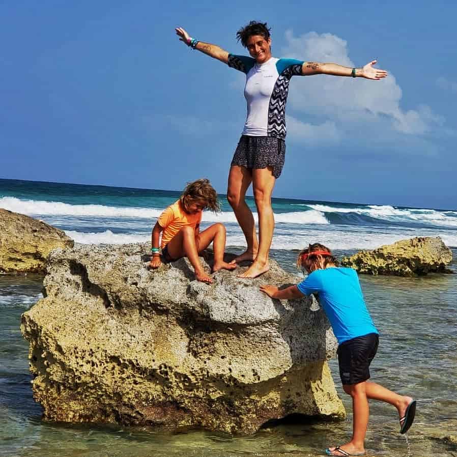Cassie in white swim shirt and two kids. Cassie standing on rock, one kid sitting at her feet and other kid about to climb up. Blue sea behind