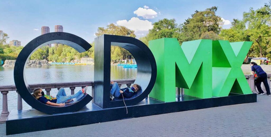CDMX letters (CD black, MX green) kid inside C and D. Blue skyand lake behind the letters