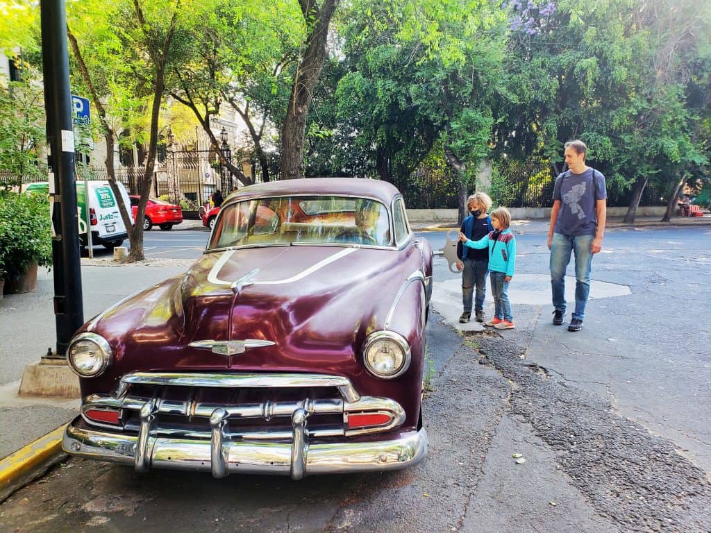 two small kids and adult man looking at an old maroon car 