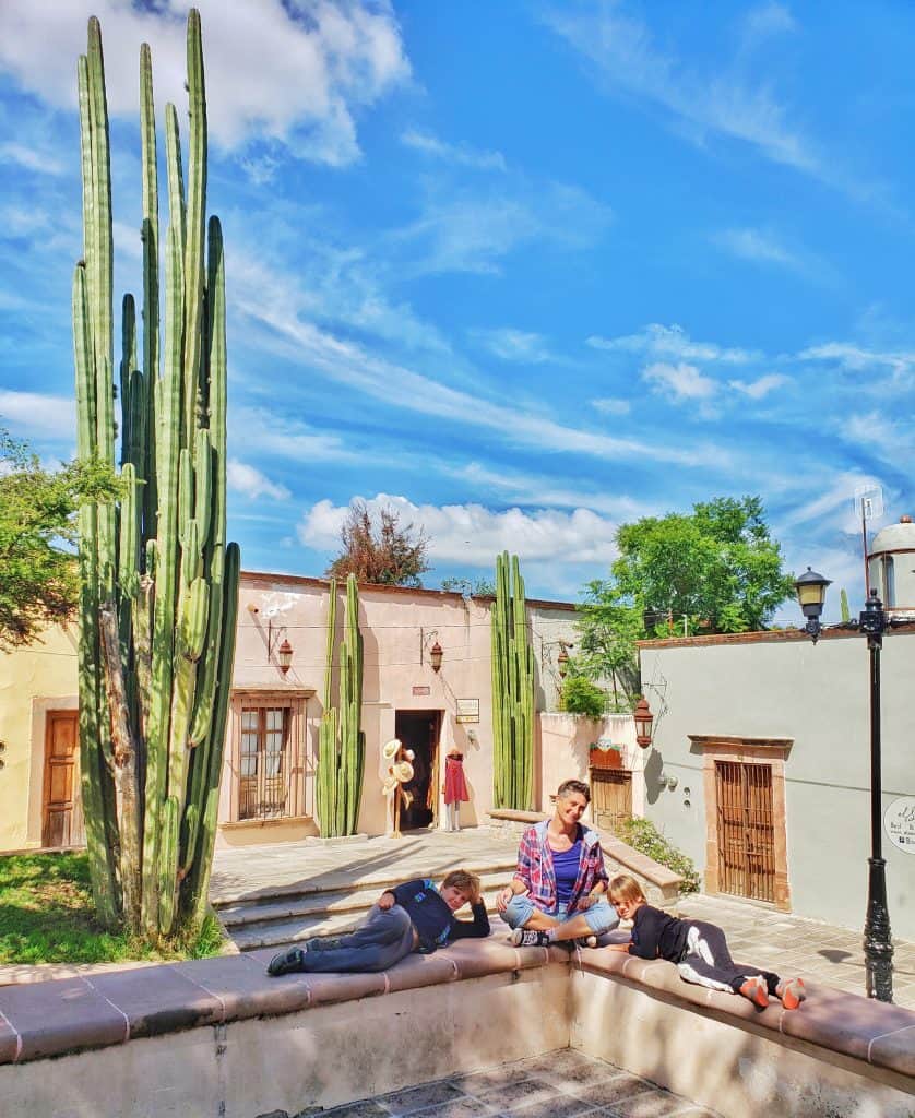 adult and two children. adult sitting crossed legs, children lying one on either side. shops and cacti in background