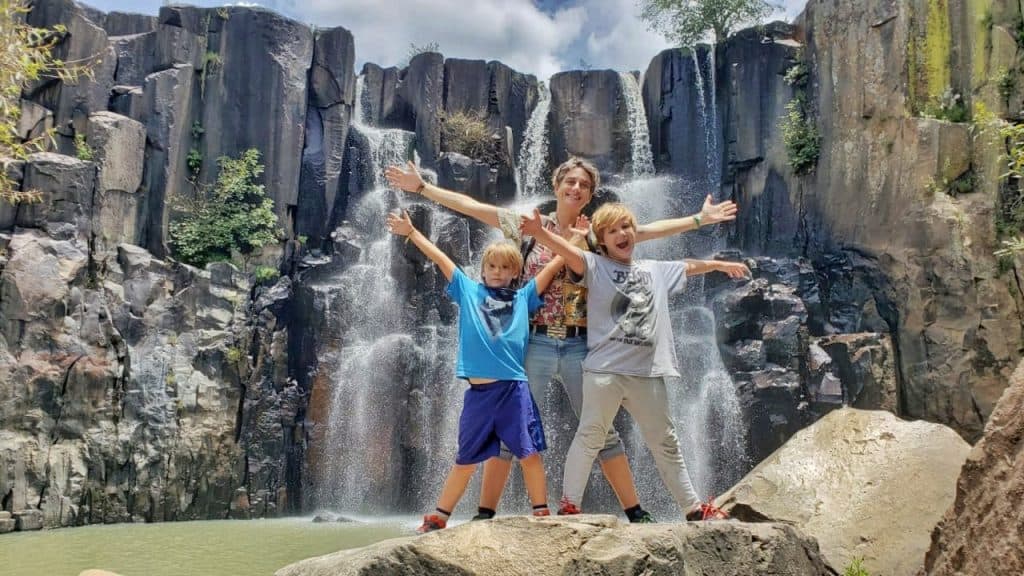 Adult and two kids standing, arms outstretched in front of waterfall