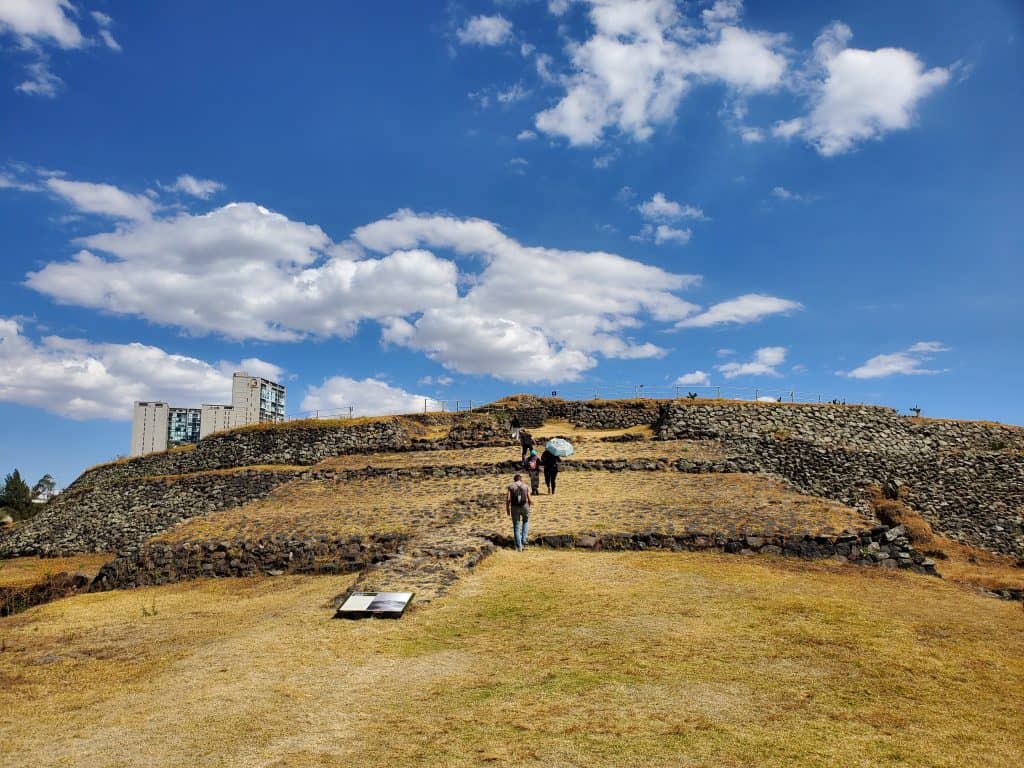 parched grass leading to ruined pyramid, people walking up, very blue sky, some clouds