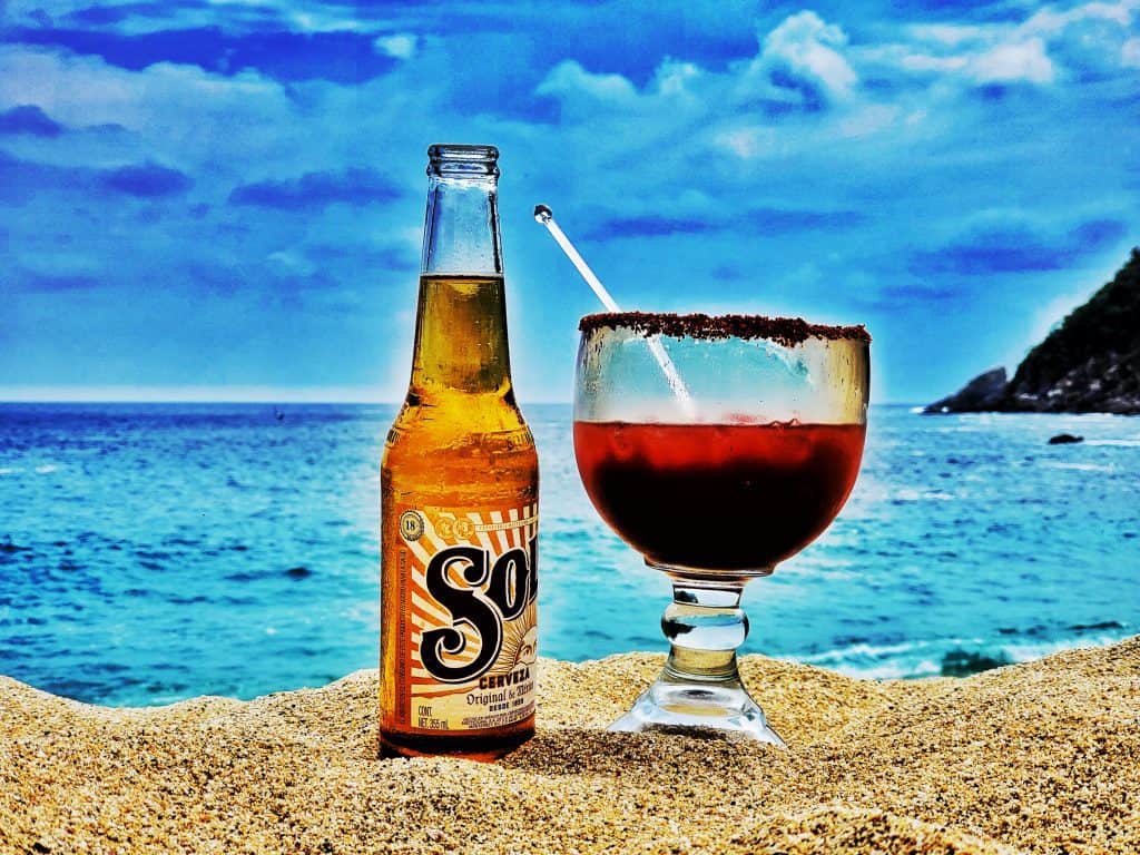 Bottle of sol beer and a glass with red michelada mix balanced in sand, ocean behind. 