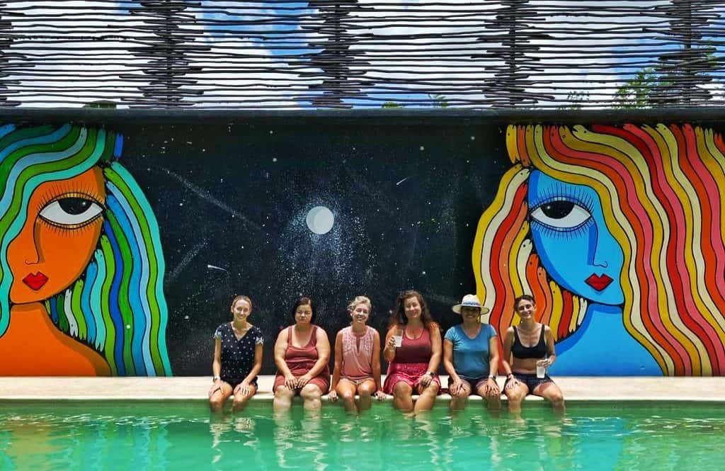 group of women sitting on edge of pool, wall behind with two mural women heads