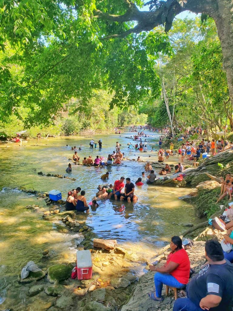 shallow river full of bathers and trees by sides
