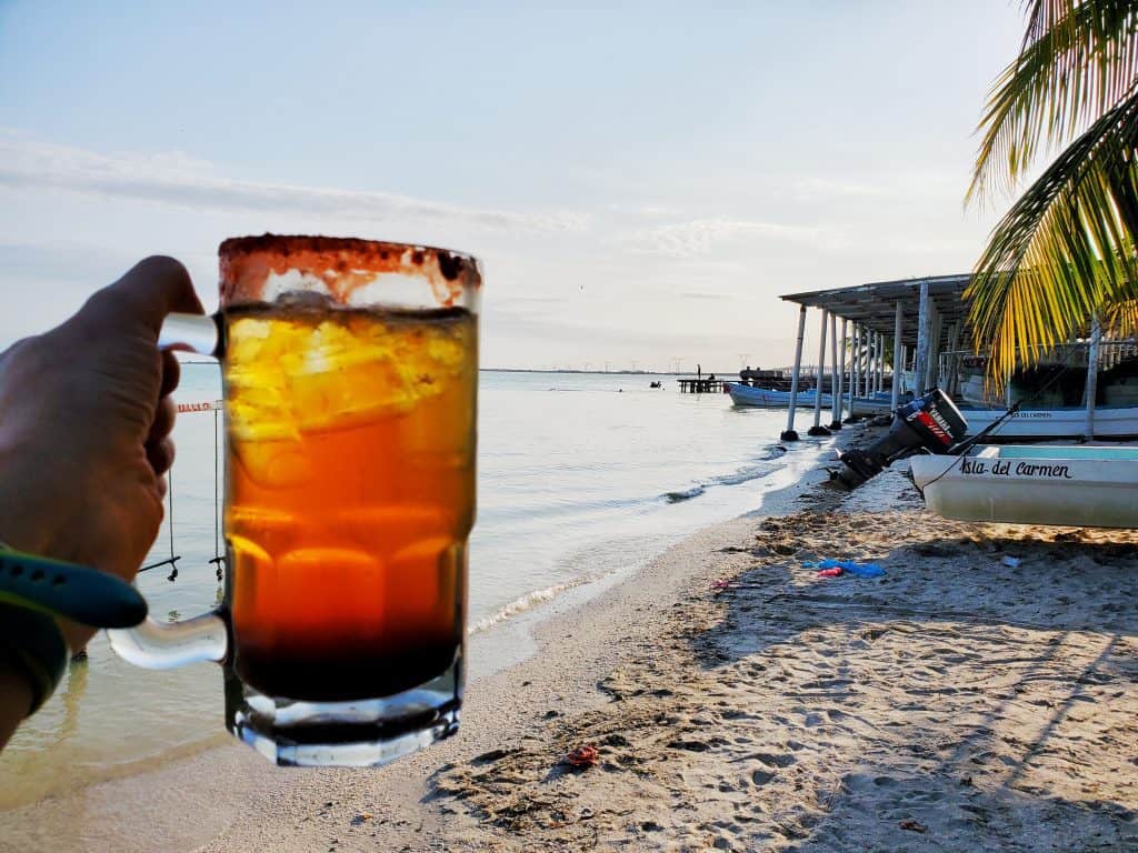 michelada held in front of pale blue sea and sand