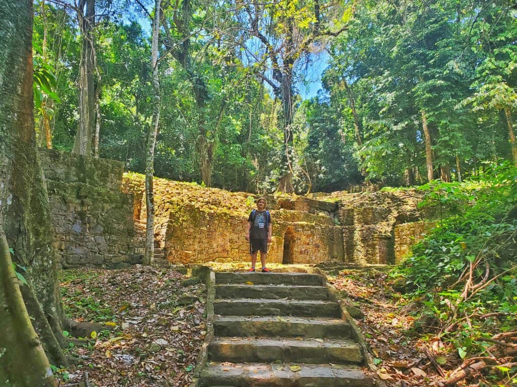 kid standing on top of small flight of steps surrounded by ruins and tall trees