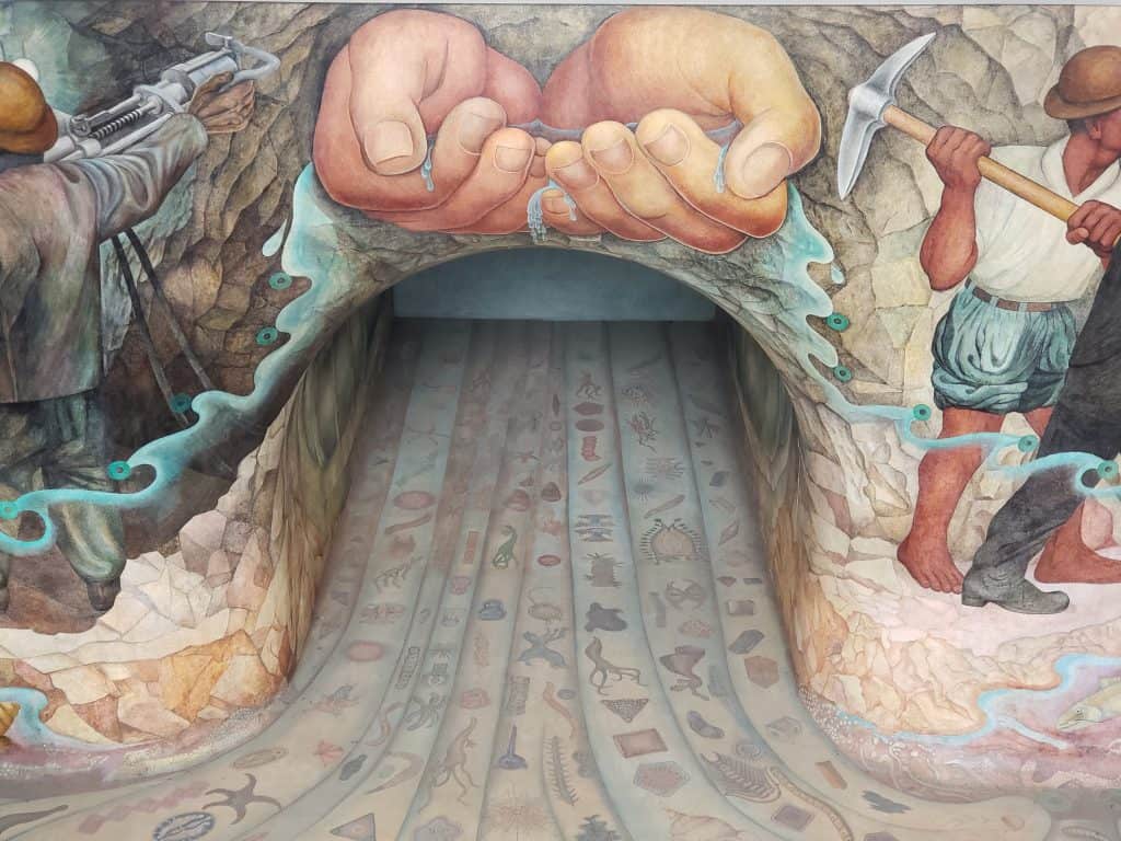 rivera mural, tunnel. floor covered in individual pictures, above tunnel, hands holding water, to right an indigenous person in shorts and white shirt with pick axe