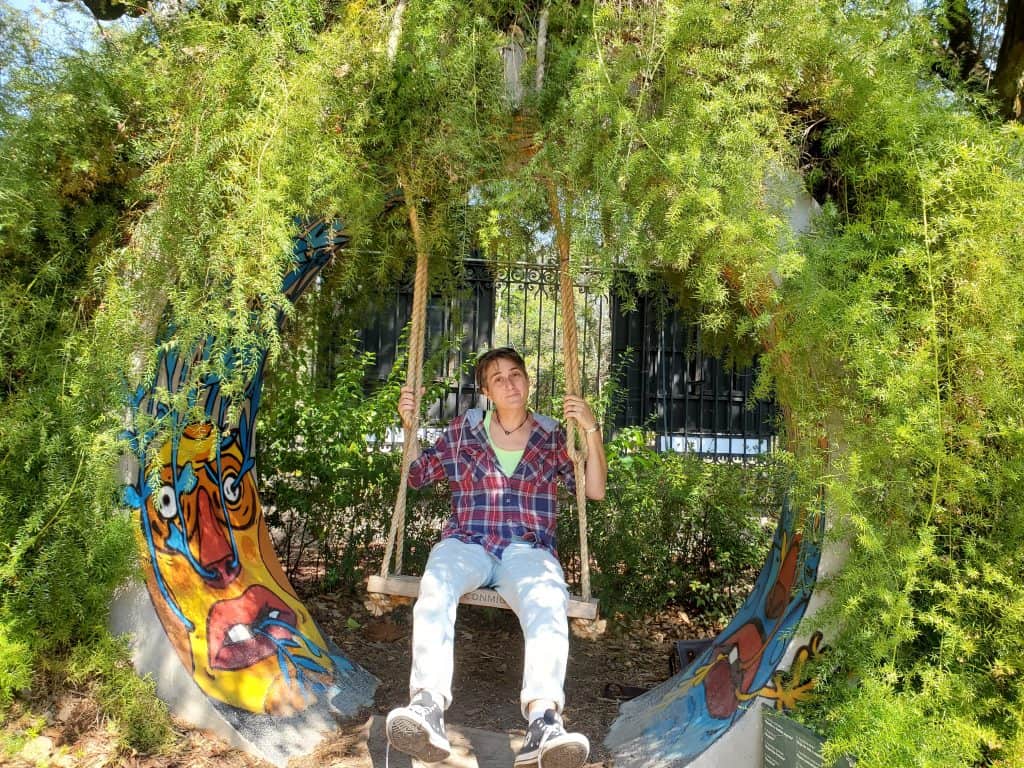 person in check shirt and jeans on a swing inside a section of concrete pipe that is covered in tree