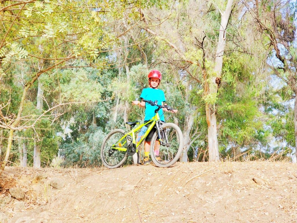 child in green top and red bike helmet standing with yellow bike in front of trees