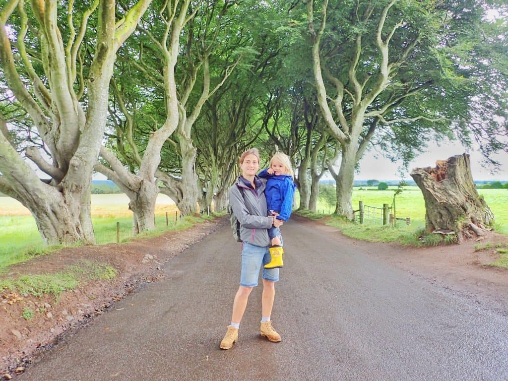 adult in grey rain coat and denim shorts holding small child in blue coat standing on country road surrounded by trees