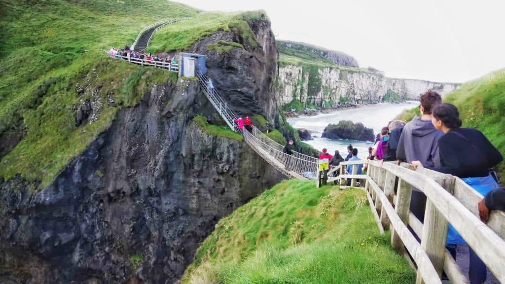 people waiting to cross a rope bridge between two cliffs