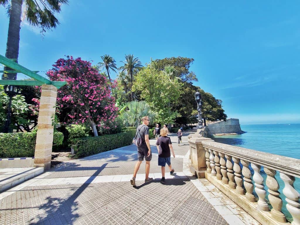 adult and child walking along sunny path, sea on one side, trees on other