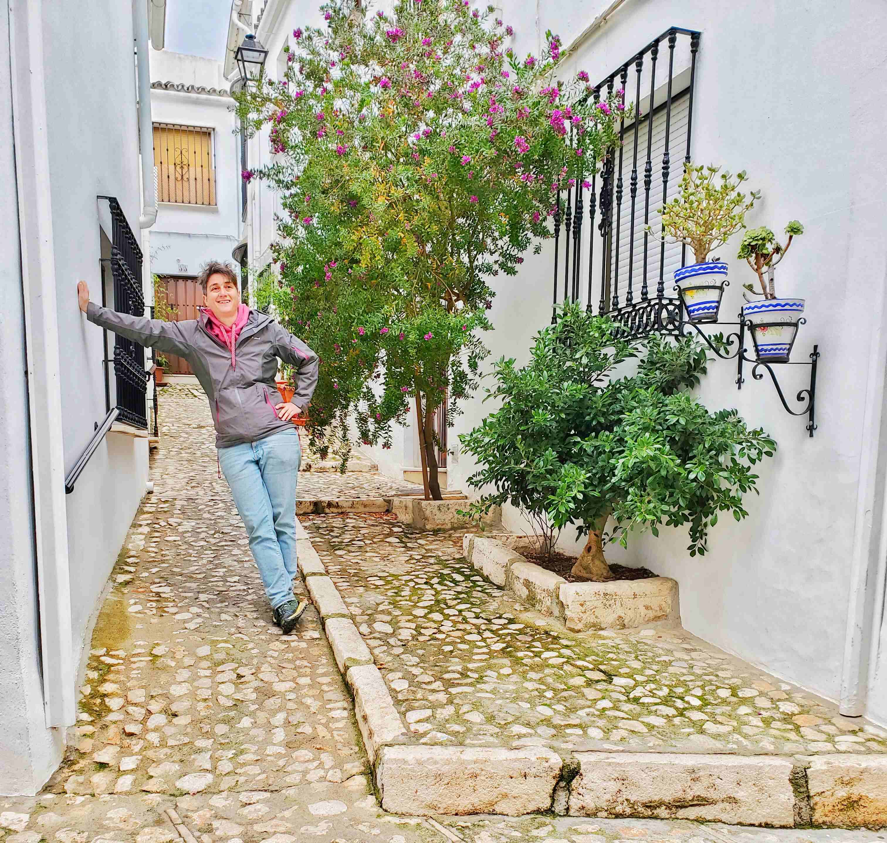 Person in jeans and grey jacket leaning on white wall in small alley way with two trees nearby