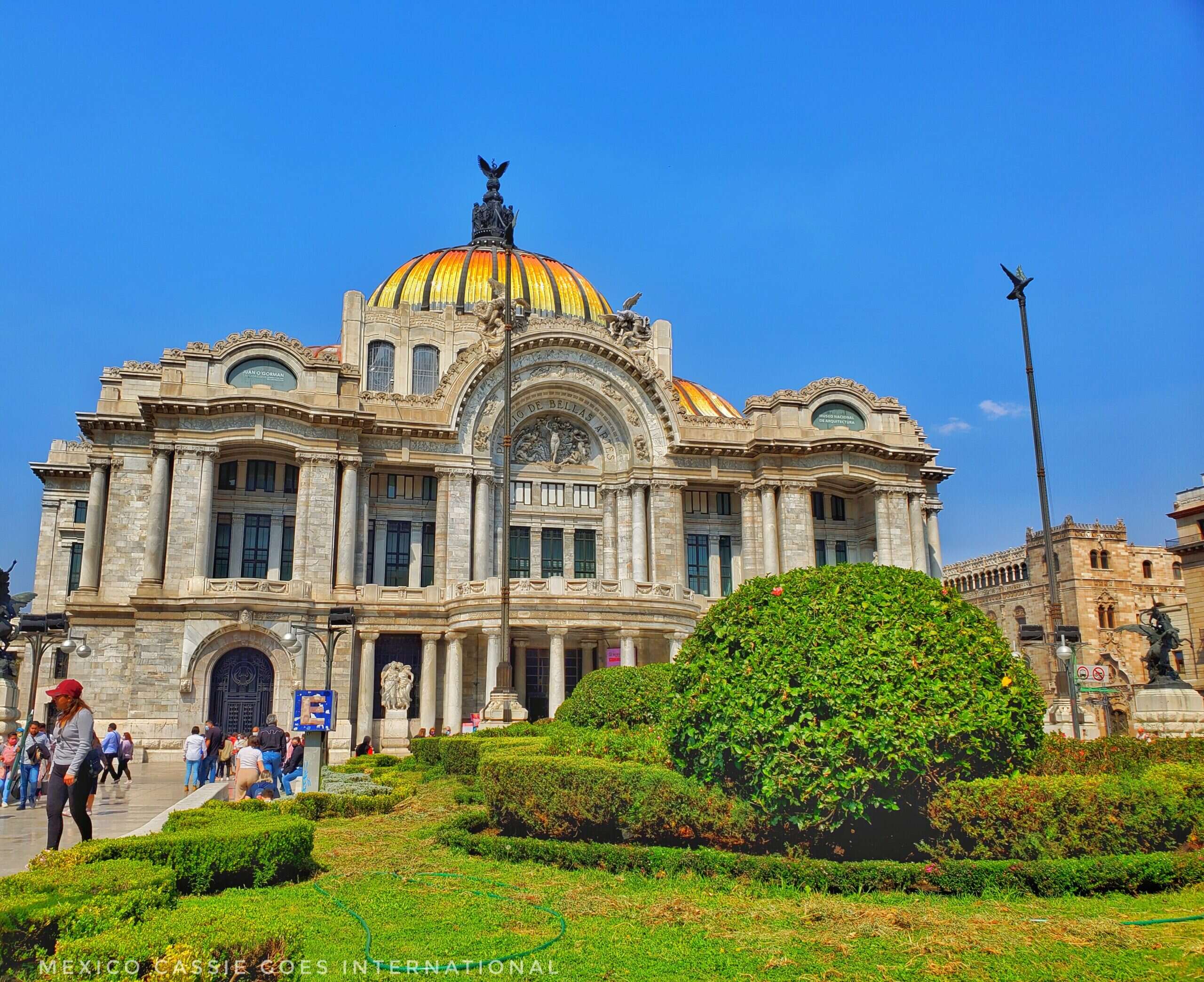 Palacio de Bellas Artes: huge building with yellow and orange domed roof. Grass and bushes in front.