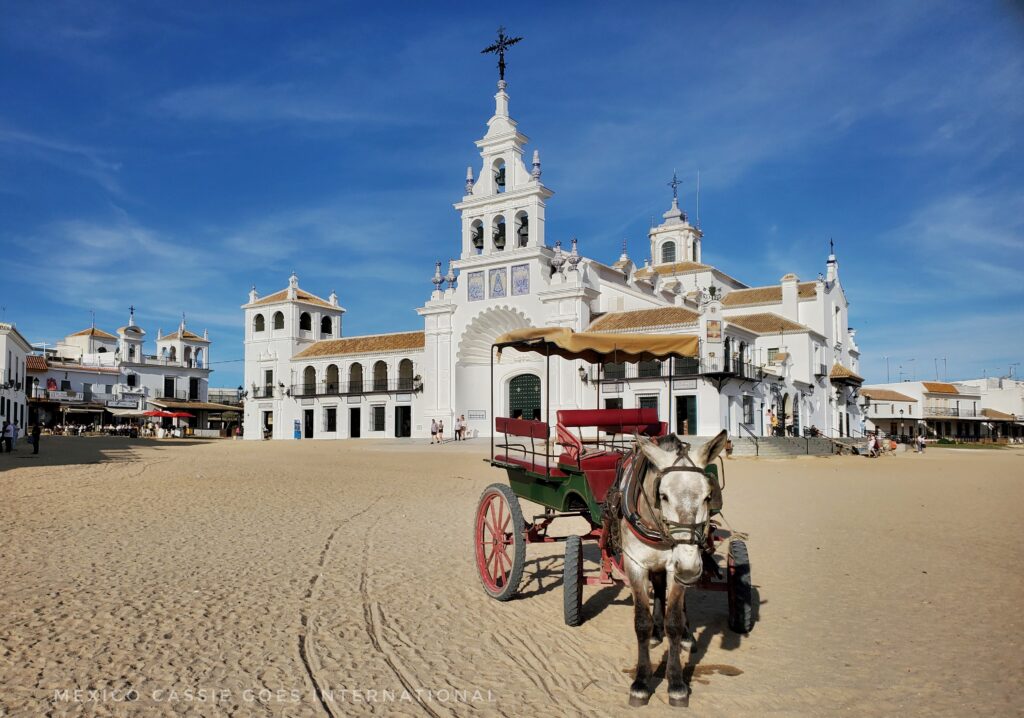 horse and cart facing camera, huge white modern church in background. sandy roads and blue sky