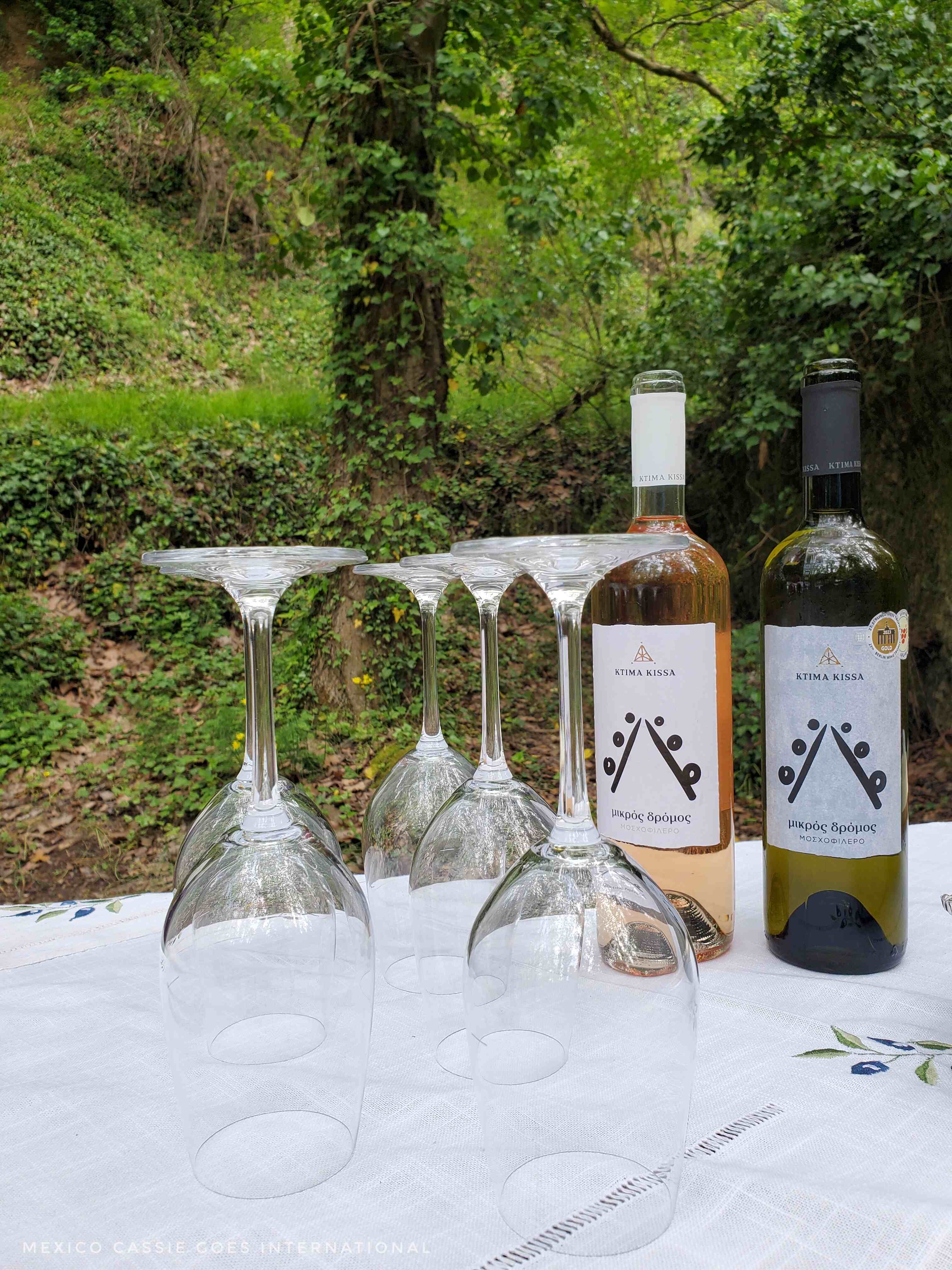 four wine glasses, upsidedown on a table next to two bottles of wine (white and rose). Backdrop is a forest