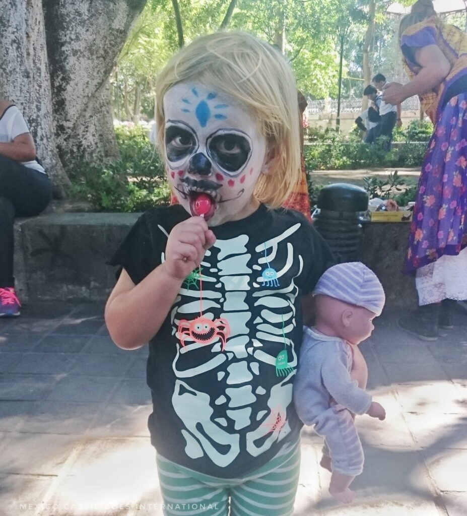 dia de los muertos 2023 - small child with face painted as a skull. holding a doll in purple. kid wearing a black skeleton tshirt and sucking a red lolly