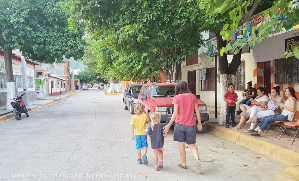 woman and two kids walking down quiet road, people on chairs on right of them