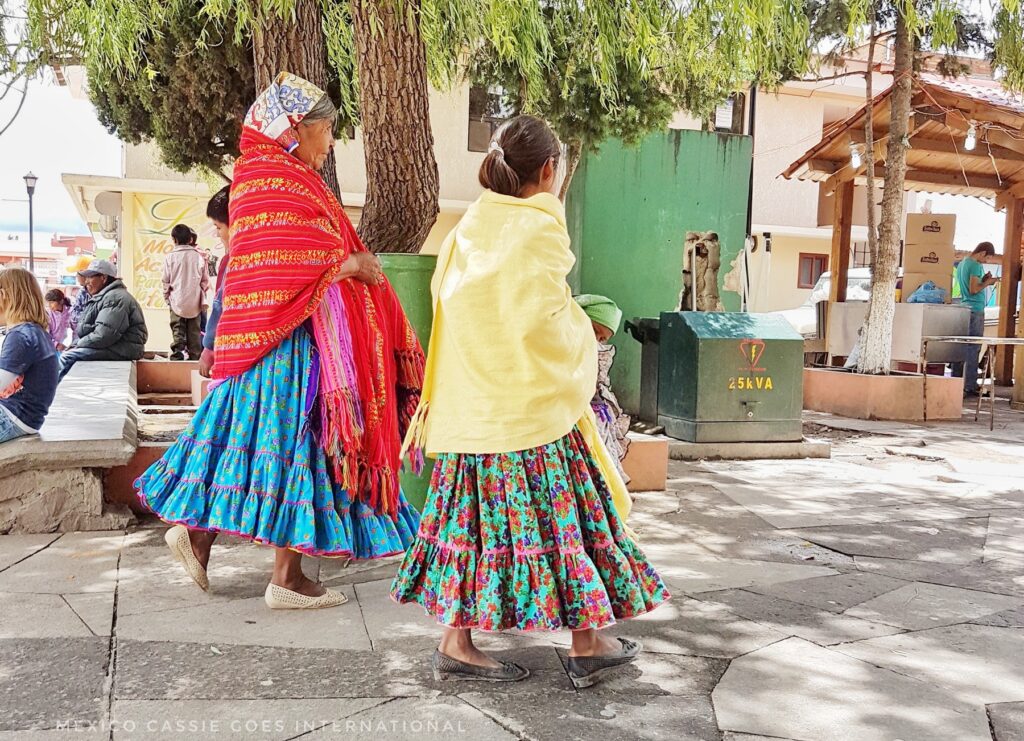 two raramuri women in creel - one in blue skirt and red scarf, other in green skirt and yellow scarf. Walking across frame.