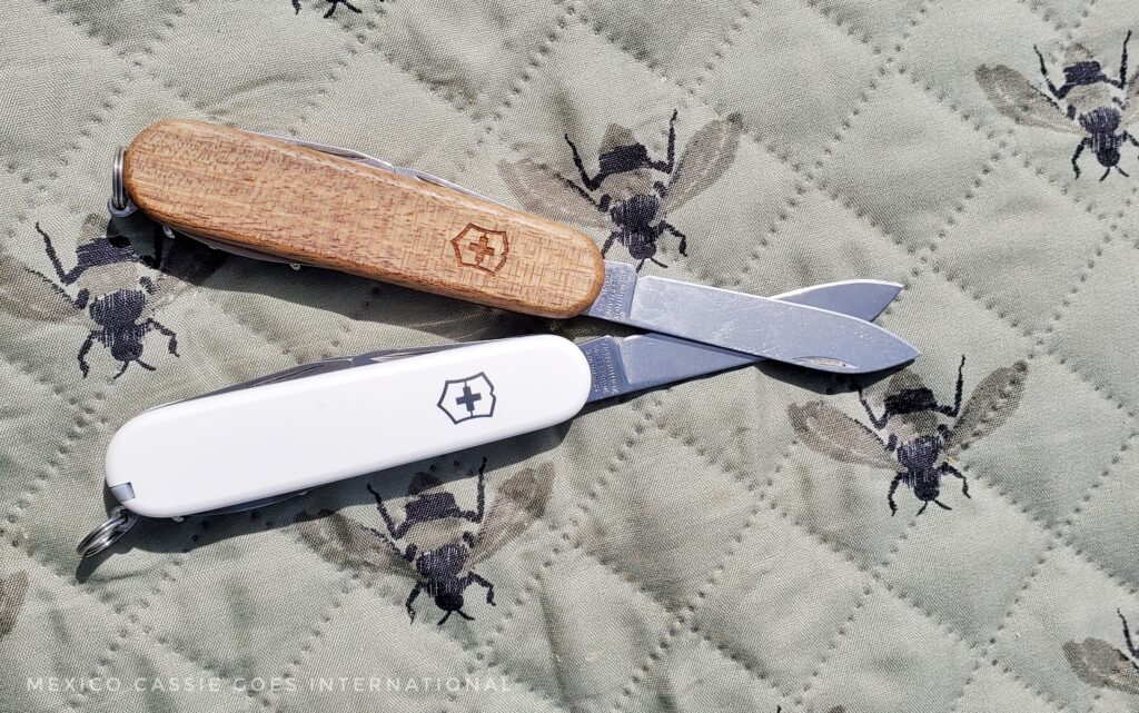 two pocket knives - one white, one wooden, knife out on each. resting on a green mat with black bees on it