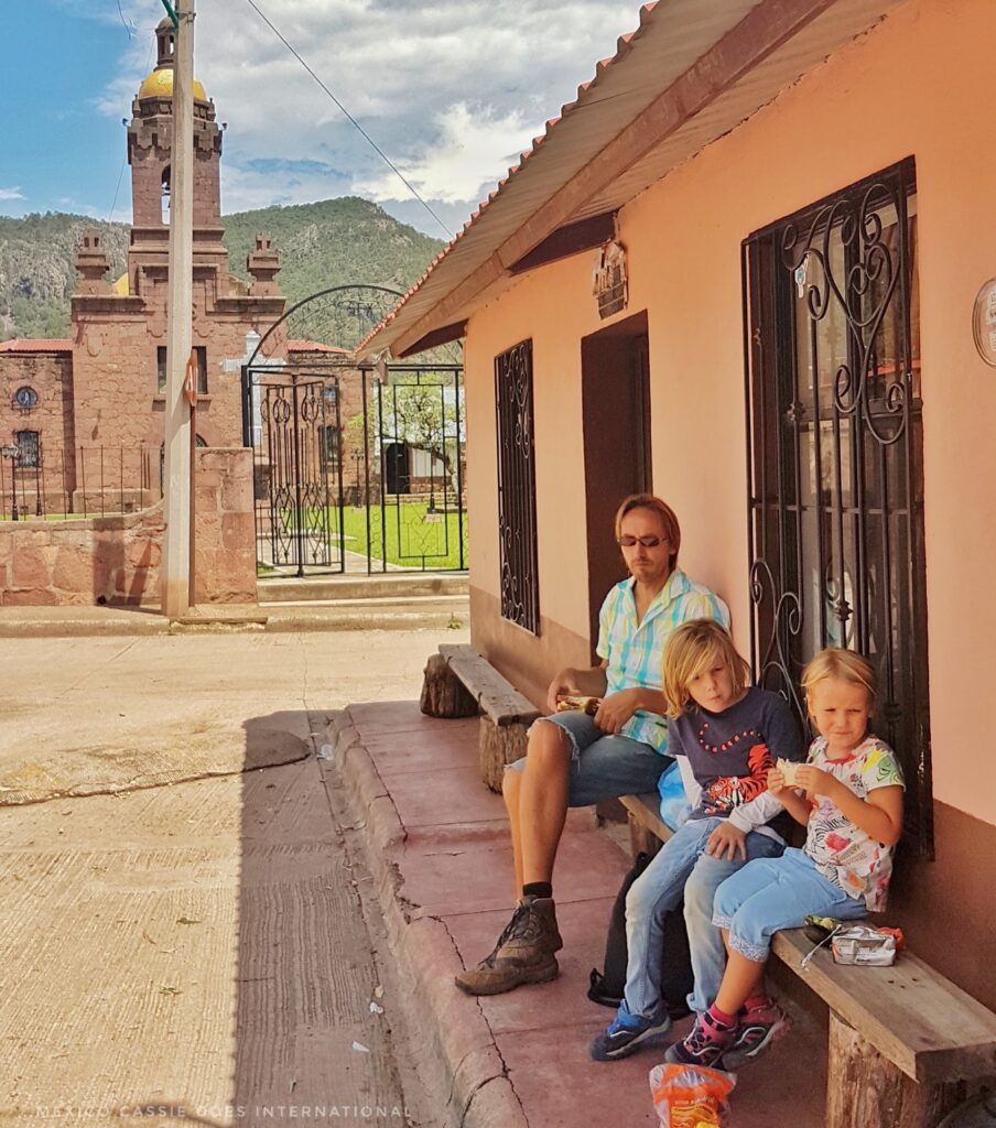 man and two children sitting on bench outside peach coloured building, church behind them