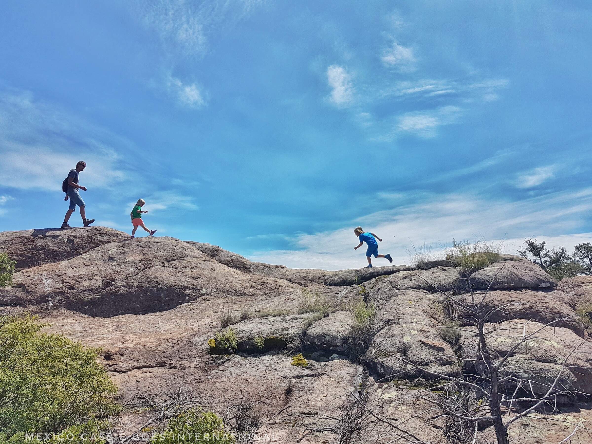 adult and two kids running along top of a rock. blue sky above