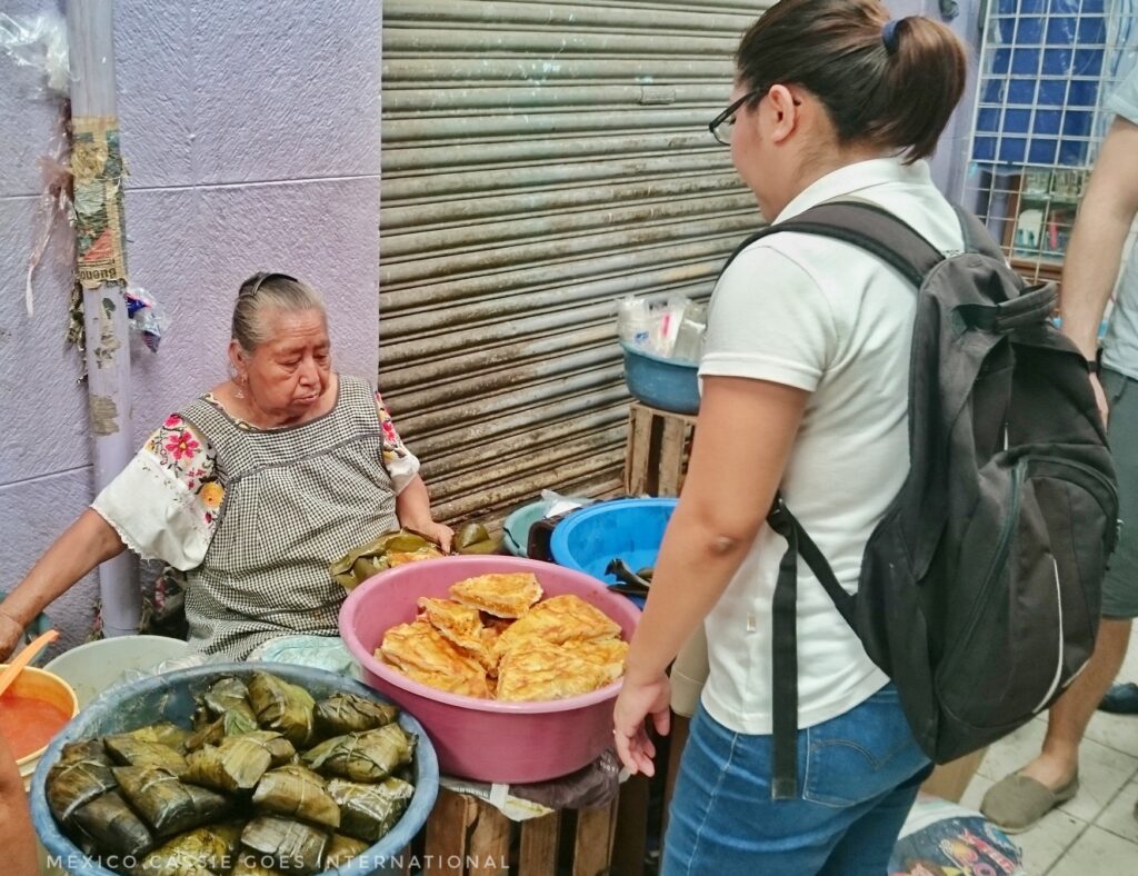 woman buying tamales from a seated woman in traditional Mayan dress. Tamales are in a washing bucket in front of her