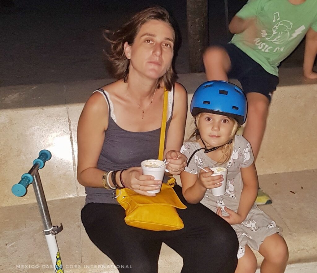 adult woman and child eat cups of corn together. woman has yellow handbag, child in blue helmet. scooter on left